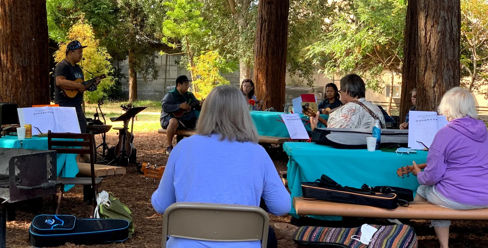 An in-person MixLife ukulele class outdoors under redwoods: the instructor plays while learners follow along at picnic tables with songbooks